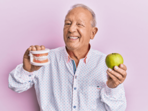 Homem idoso segurando uma maçã e uma prótese dentária